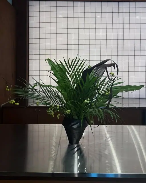 격자 유리창을 등지고 스테인리스 카운터에 놓인 초록 식물 화병. 창으로 들어온 빛이 카운터에 반사된다.