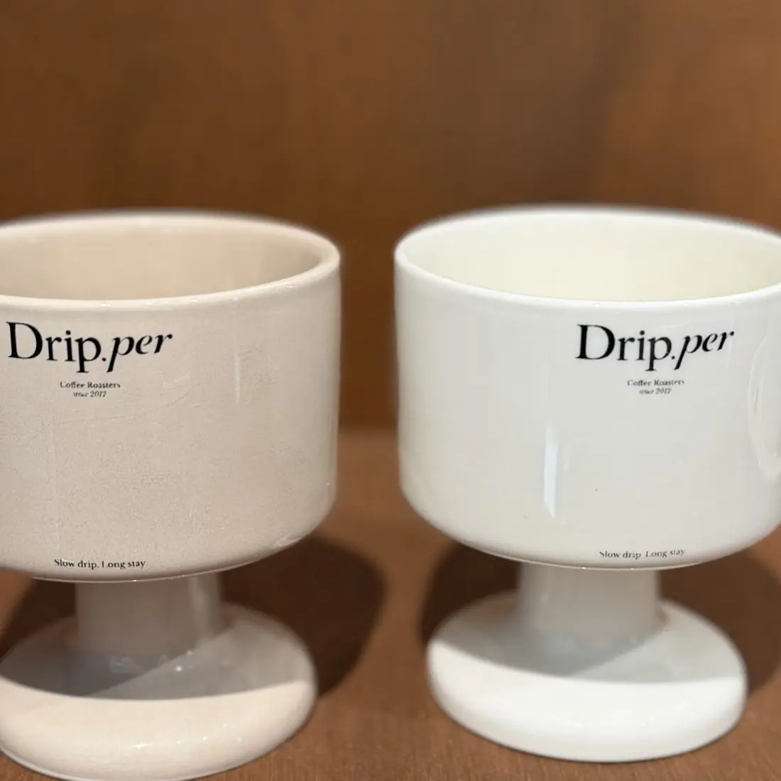 우드 선반에 나란히 놓인 Drip.per 굽 잔 두 개. 크림색과 흰색이며 Slow drip. Long stay. 가 적혀 있다.