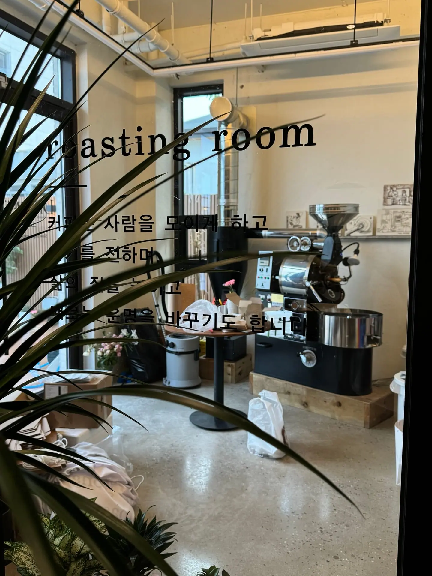 유리문 너머로 보이는 로스팅룸. 문에는 roasting room 이라는 글자와 커피에 관한 문장이 새겨져 있고, 안쪽에 로스터기와 천장으로 올라가는 배기 덕트가 보인다. 문 앞으로 큰 관엽식물의 잎이 드리워져 있다.